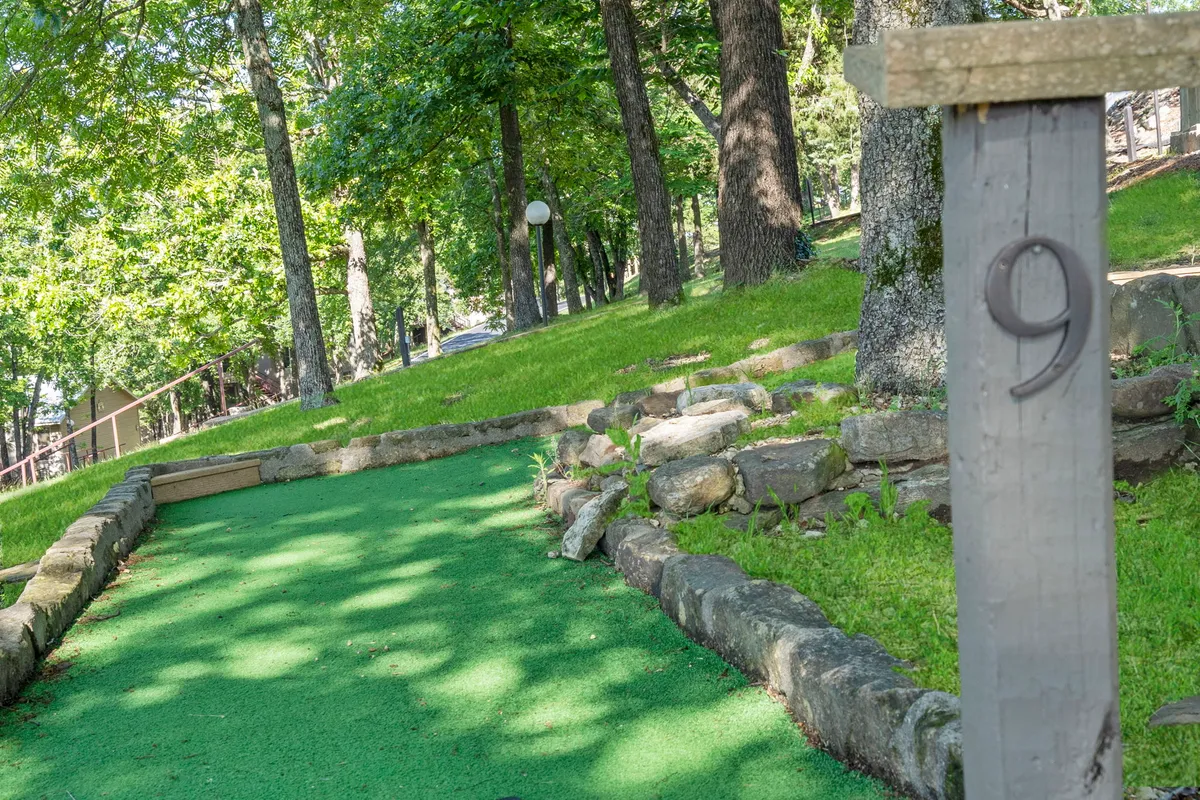 A vibrant mini-golf putting green bordered by stones, with a numbered post and surrounded by tall, leafy trees. The space looks inviting for families, with clear areas for gatherings and play.