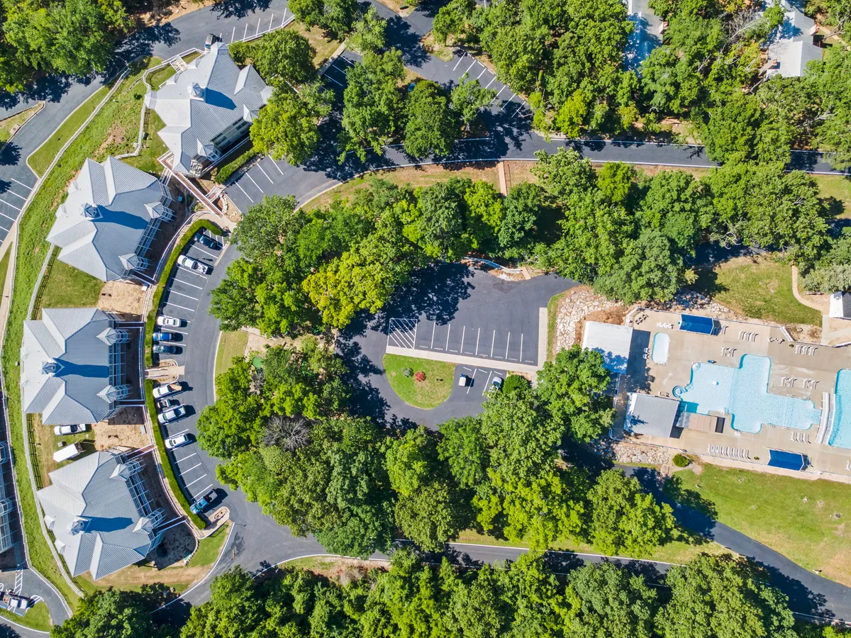 A relaxed view of residential buildings flanked by a swimming pool, with guests lounging and enjoying sunny afternoons. Trees offer shade along the walking paths and the atmosphere is peaceful yet lively.