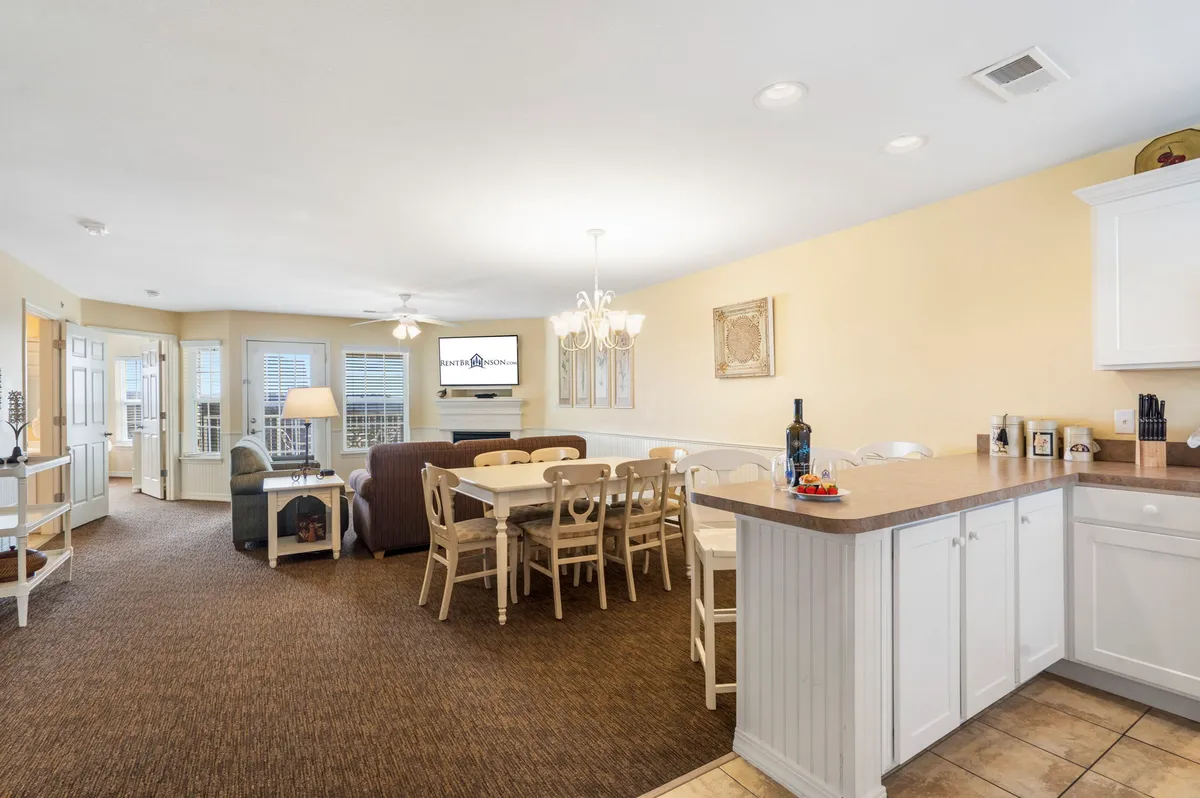 An open-concept space with a dining table, kitchen island, sofa, chandelier, and a television in the background. The arrangement creates a seamless flow between living, dining, and kitchen areas, with warm lighting and inviting decor.