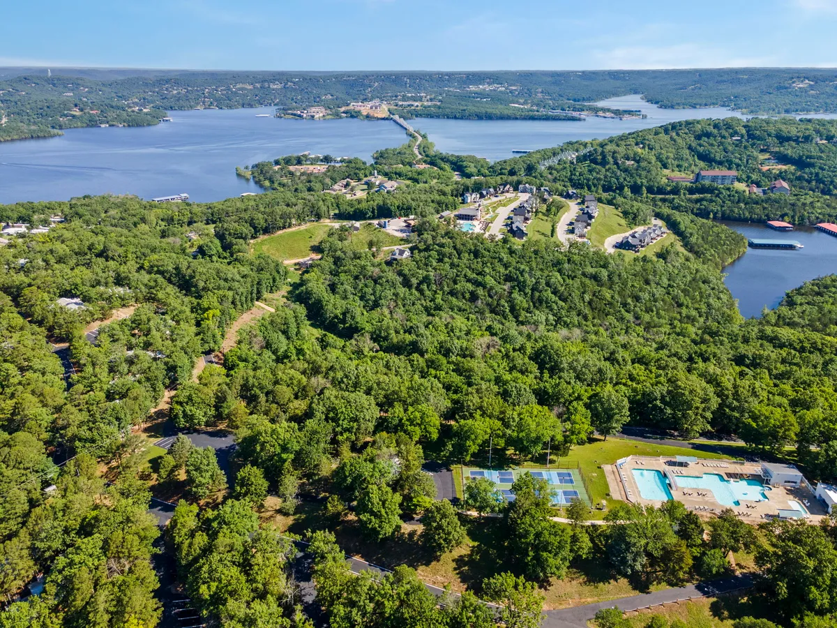An elevated view showcasing lush greenery, a swimming pool, tennis courts, and the sparkling lake beyond. Residential buildings are nestled into the landscape, emphasizing the blend of natural beauty and resort-style amenities available to guests.