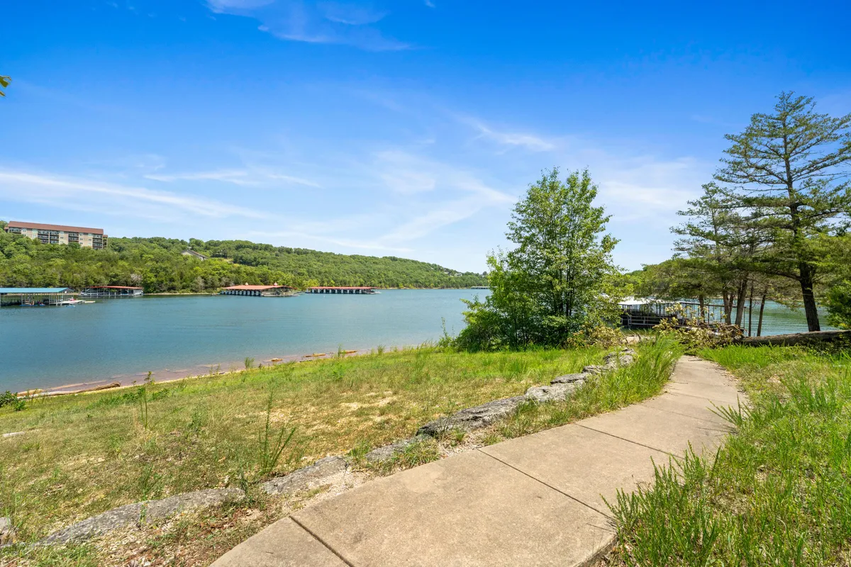 A tranquil riverside scene featuring a winding pathway lined with trees, gentle hills in the background, and calm water glistening in the soft light. The setting invites leisurely walks and moments of serenity amidst nature.