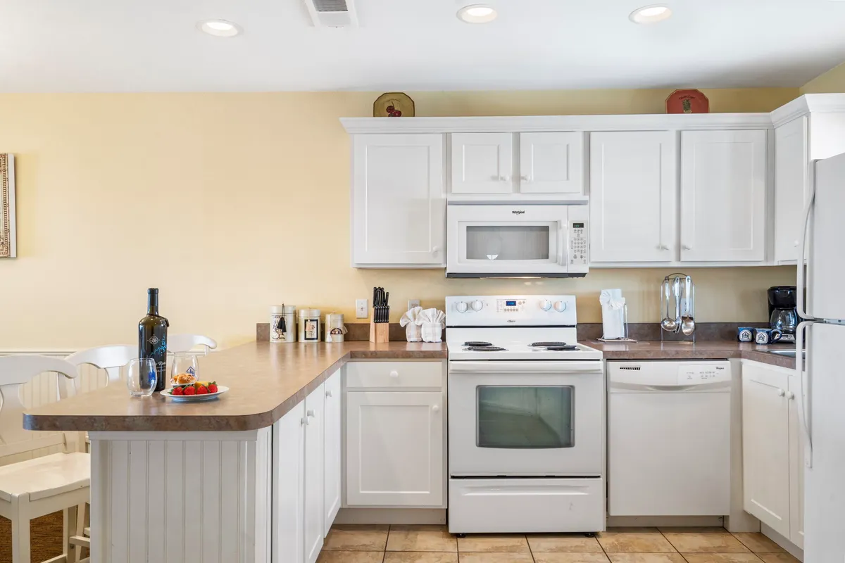 A modern, fully equipped kitchen with a spacious island, white cabinetry, stainless steel appliances, a bottle of wine, and a bowl of fresh fruit. The layout and amenities cater to cooking and entertaining.