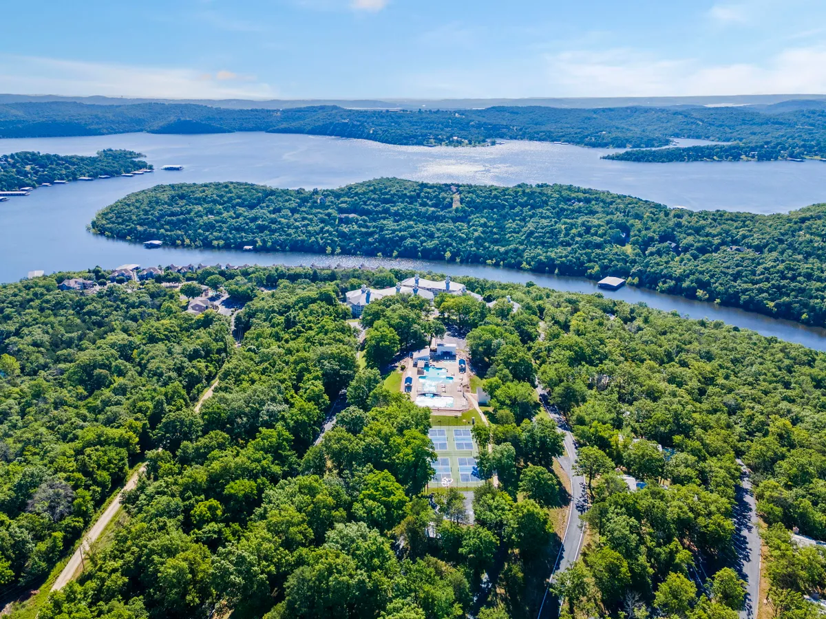 This image captures an expansive outdoor setting with water views, lush greenery, a swimming pool area, and tennis courts. There are people enjoying various activities such as swimming, tennis, hiking, and relaxing by the lake, all set against a tranquil, recreational backdrop.