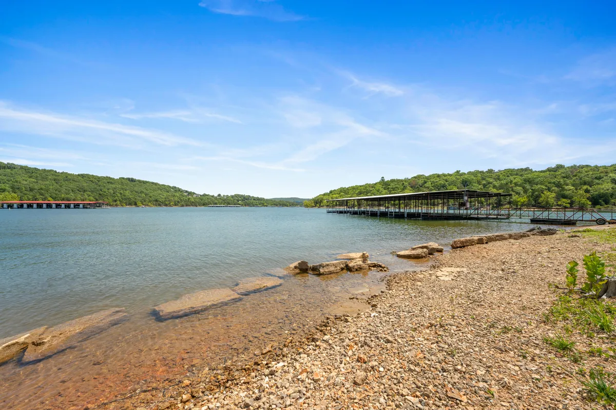A peaceful lakeside scene with calm waters, docks stretching out over the lake, a rocky shoreline, and green hills in the background. The environment suggests opportunities for boating, fishing, and nature walks along the shore.