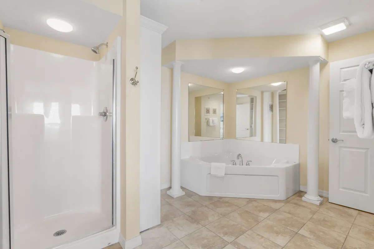 A luxurious ensuite bathroom featuring a jacuzzi tub, separate shower stall, elegant columns, and mirrors. The decor is calming, designed for spa-like pampering and relaxation.