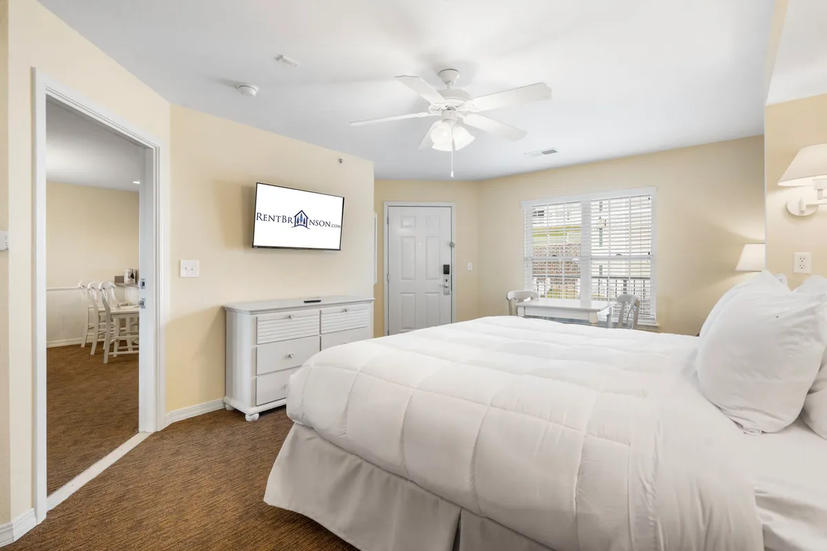 A bright, comfortable bedroom with a king-sized bed, television, and abundant natural light streaming in from large windows. The setting is tranquil and designed for restful sleep or quiet moments.