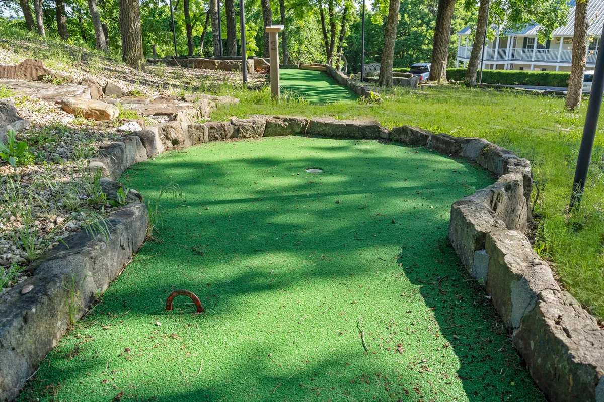 A cheerful mini-golf putting green bordered by stones and surrounded by tall trees. Friends and families are gathered, some playing mini-golf while others enjoy a picnic or relax nearby, fostering a fun and relaxed mood.