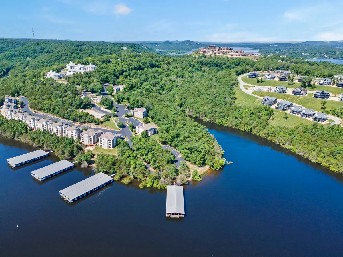 The image presents the exterior of residential buildings nestled near a lake, with visible docks, hills, and a road leading to nearby trails. The setting feels peaceful and naturally integrated with its surroundings.